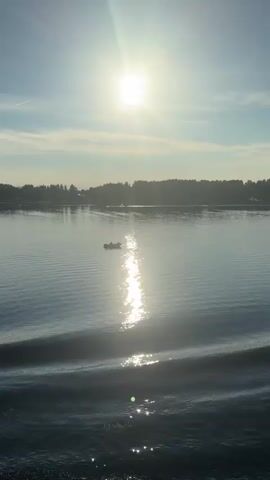 The video shows a body of water with a boat in the distance. The sun is high in the sky, casting a reflection on the water. There are trees on the horizon. The water is calm with small waves.
