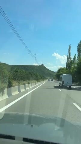 The video shows a first-person perspective from inside a moving vehicle on a multi-lane highway. The sky is bright blue with scattered white clouds. Power lines stretch across the top of the frame, supported by utility poles. On the left side of the highway, there is a concrete barrier and a hillside covered in green trees. In the distance, a structure, possibly a castle or fortress, is visible on top of a hill. On the right side of the highway, a white van is driving parallel to the camera. More green trees and a clearer view of the distant hills are visible on the right. The road ahead appears to be clear, with other vehicles in the distance. The dashboard of the vehicle is visible at the bottom of the frame.