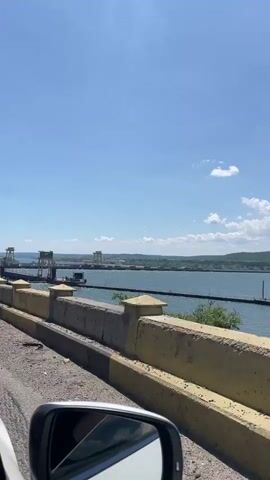 The video shows a bright sunny day with a clear blue sky, a car driving along a road next to a body of water. The road has a concrete barrier on the right, and then a metal railing. Beyond the railing is a wide expanse of water with several large structures, possibly cranes or parts of a dam, in the distance. Hills are visible on the far side of the water. The car's side mirror is visible in the bottom left of the frame. At 0:00, a blue road sign with white text that reads "Padina Crucii" appears briefly on the left side of the frame.