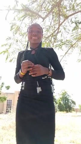 A young woman in a black long-sleeved dress dances under a tree on a sunny day. She has her hair in braids, and wears gold bracelets. She is smiling and facing the camera, then turns slightly to her right. A building is visible in the background to the left.