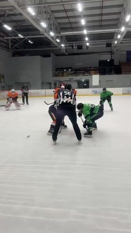 A hockey game is in progress on an ice rink. The scoreboard shows a score of 2-3 with approximately 11:52 left in the period. Two players in green uniforms are on the ice, one in the foreground with a stick in hand, and another further back in the mid-ground. Two players in orange uniforms are also on the ice, one in the foreground near the net and another in the mid-ground. A referee in a black and white striped jersey is positioned between the two green-uniformed players in the foreground. An orange-clad goaltender is in the net on the left side of the frame. The rink has white ice with red and blue markings. The environment appears to be an indoor arena with a high ceiling, exposed metal beams, and fluorescent lighting. The video is shot from a low angle, close to the ice surface.