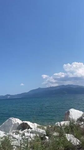 A bright, clear day with a vast blue sky stretches above a calm turquoise sea. In the foreground, a rocky shoreline is dotted with white rocks and green foliage. Across the water, a range of distant mountains forms a hazy silhouette against the horizon, with a few scattered clouds drifting in the sky. The scene evokes a sense of peaceful, natural beauty.