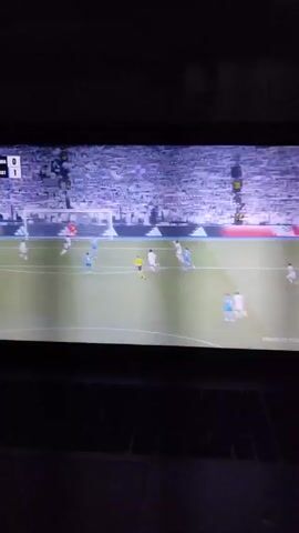A TV screen shows a soccer game with players wearing white and light blue uniforms on a green field. The players are positioned around the penalty box in front of a goal. The score on the screen shows “0-1”. A large crowd wearing white fills the stands behind the goal. The screen is dark except for the bright image of the soccer game. The surface on which the TV sits appears to be a dark-colored laptop.