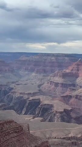 A sweeping aerial view of the Grand Canyon. The vast, layered rock formations stretch as far as the eye can see. The colors range from deep reds and purples to muted browns and grays. The sky above is overcast with dramatic, textured clouds. The scale of the canyon is immense, emphasizing the rugged terrain and the vastness of nature. There are no people or man-made objects visible. The video provides a sense of depth and grandeur, showcasing the geological wonder of the canyon.