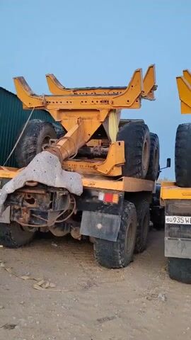 This video shows the rear view of two large yellow industrial vehicles, likely trucks or specialized machinery, parked next to each other outdoors on a dirt or gravel surface. The sky is visible in the background. The vehicles have large, thick tires. The vehicle on the left has a wooden plank secured to its chassis, supporting part of the yellow equipment attached to it, which appears to be some sort of lifting or gripping mechanism with large, curved arms. A white, patterned cloth or cover is draped over a part of this mechanism. The vehicle on the right also has a similar yellow attachment on its rear. Its license plate is partially visible and reads "BG 935 WBA". The overall impression is that these are heavy-duty vehicles, possibly in storage or awaiting transport or work.