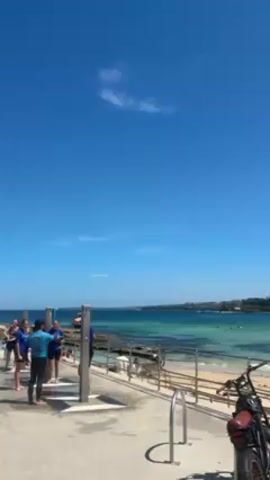 The video shows a group of people standing near outdoor showers on a sunny day. The people are near a beach with clear blue-green water. A sandy beach and some greenery are visible in the background. The people appear to be washing off after swimming or surfing. One man wearing a blue shirt walks by the group. A motorcycle is parked on the edge of the frame. The sky is clear blue with a single, small white cloud.