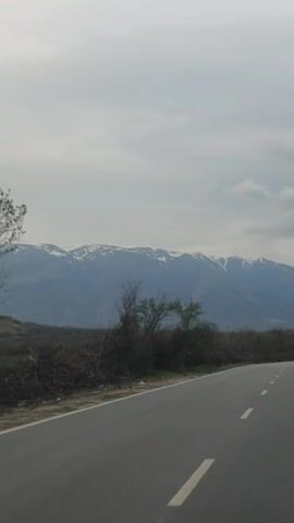 The video depicts a gray asphalt road with white painted stripes leading towards a distant mountain range with snow-capped peaks, all under a cloudy sky. Sparse vegetation lines the road, including a few bare trees and low shrubbery. A single vehicle is visible far down the road, approaching the viewer.