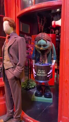 A still mannequin dressed in a pinstripe suit and tie stands to the left of the frame. To the right, a cartoonish, green-faced figure with pigtails is depicted vomiting purple candies into a bucket labeled "Weasleys' Wizard Wheezes Puking Pastilles." The backdrop appears to be a shop interior with red architectural elements, shelves filled with various items, and decorative elements. The lighting is dim, with a warm, reddish glow emanating from the environment.