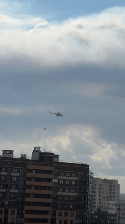 Helicopter tows red bucket across cloudy sky in aerial operation