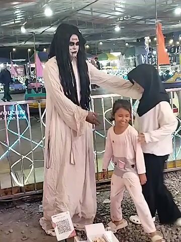 In a night scene at what appears to be an amusement park or fairground in Sragen, Indonesia, a person dressed as a ghost poses for a photo with an adult female and a young child. The ghost costume features a long black wig, white face paint with black eye and mouth accents, and a flowing white robe. The ghost, initially with an arm around the adult female, smiles broadly while posing. The adult female, wearing a black hijab, white top, and black pants, smiles throughout. A young child, dressed in light-colored pajamas, runs playfully near them, laughing at one point, and also smiles during the pose. In the background, bumper cars are visible, along with metal fences and various fairground lights, suggesting an active area. The ground is covered with gravel or dirt. On the ground in front of the ghost, a small box containing items and a sign displaying a QR code are visible. A partial sign with the word "Voucher" can also be seen in the background. A voice is heard counting to three, indicating that a photo is being taken.
