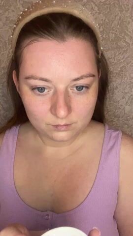 The video opens with a close-up shot of a young woman with fair skin, light brown hair pulled back by a sparkly, light-colored headband, and blue eyes. She is wearing a light purple ribbed tank top. Her complexion shows some freckles and minor blemishes. Initially, she is looking down, seemingly at something she is holding in her hand.

At the 0:00 mark, she looks up briefly, her blue eyes directly at the camera, then looks down again. She is holding a small, light-colored container, possibly a jar of cream, in her left hand, but it's mostly out of frame.

At 0:01, she dots a small amount of white cream onto her forehead and then onto her right cheekbone using her right index finger, which has light purple nail polish. She then looks down at the container again.

By 0:02, she has dabbed another dot of cream onto her left cheekbone. She continues to look down as she applies these dots.

At 0:03, she looks up again, making eye contact with the camera. She then uses her right hand to gently spread the cream onto her forehead, moving in small, circular motions. Her expression is neutral, focused on the task.

The video is filmed from a slightly elevated angle, looking down at the woman. The background is a textured, light brown or beige wall, appearing indoors. The lighting is soft and even, illuminating her face clearly. The focus is entirely on her and her actions of applying the facial product.