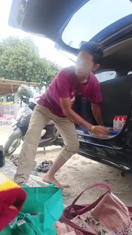 The video shows a young man in a purple t-shirt and tan pants standing next to the open trunk of a black car. He is barefoot and has a bag slung across his shoulder. He is reaching into the trunk, and a red duffel bag is being handed to him from the left. In the trunk, there are several cylindrical objects stacked, and a blue mat. In the foreground, there are several bags, including a green tote bag and a patterned bag. In the background, there is a motorcycle, a building, and trees. The overall scene appears to be outdoors in daylight.