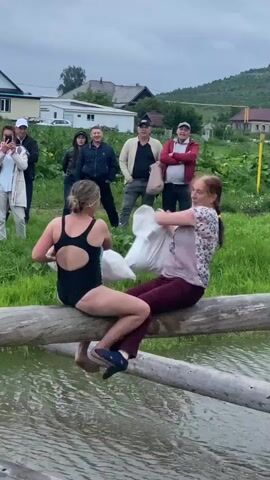 Two women are seated on a log that is positioned over water, engaged in a pillow fight. The woman on the left is wearing a black swimsuit and the woman on the right is wearing a floral shirt and maroon pants. They are both holding large white pillows and striking each other with them. Several people are watching them from the grassy bank behind the log. The environment appears to be a rural outdoor setting with houses and a hill in the background. The sky is overcast.