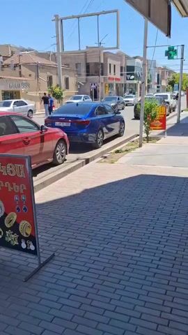 The video shows a street scene on a sunny day. Several cars are parked along the street, including a red sedan, a blue sedan, and a white SUV. A red and white sign displaying jewelry is on the sidewalk. There are buildings in the background with shops and signs. People are visible walking on the sidewalk and standing near the cars. A tree is planted on the sidewalk. The street is paved with brick.