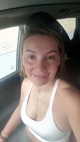 A woman in a white tank top sits in the driver's seat of a car. She smiles at the camera, looks to her right, and then makes a peace sign. She has sunglasses on her head and is wearing bracelets and a necklace. Latin music is playing in the background. The car is parked and the windows are up.