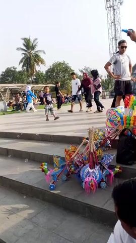 The video shows an outdoor scene, possibly a park or public square. The foreground is dominated by a set of concrete steps. On the steps, a collection of colorful toys is displayed, including what appear to be wooden toy horses with elaborate designs and large balloons. Some of the balloons are orange and yellow, others have cartoonish faces. A black plastic bag full of items, possibly more toys or balloons, sits near the toys. Several people are visible in the midground and background. On the steps, a man in a light-colored t-shirt and blue capri pants, wearing flip-flops, stands near the toys, looking down at them. Another man, wearing a white t-shirt and sunglasses, stands with his hands on his hips in the background, to the right. Further back, a group of people, including a man in a white t-shirt and shorts, a woman in a pink hijab and a woman in a dark top, are walking. A young boy in a blue t-shirt and shorts is standing on the steps, facing away from the camera. In the far background, there are trees, a tall metal structure, and buildings. The sky is bright and appears to be a clear day. The overall impression is a lively outdoor market or gathering place where toys are being sold.