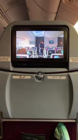Here's a description of the video:

The video shows the back of an airplane seat. The seat has a small entertainment screen displaying an airline safety video. The safety video ends, and the screen transitions to a blue screen displaying the LATAM Airlines logo. There is a small shelf below the screen with a pamphlet and a water bottle. The lighting in the cabin is bright.
