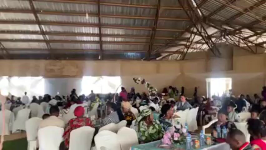 The video shows a large room filled with people seated at tables. The room has a high, corrugated metal roof supported by wooden beams. The walls are painted in a light color.  The tables are covered with white tablecloths, and the chairs are also white. Many of the people seated at the tables are dressed in colorful clothing. There are floral arrangements on the tables. A floral arch is visible in the background.