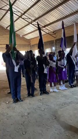 In an indoor setting with a corrugated metal roof and exposed brick walls, a group of individuals stands in a line. The individuals are holding flags and what appear to be rifles. The person on the far left is holding a flag with green and white stripes, which resembles the Nigerian flag. To their right, another person holds a black flag. The third person from the left holds a flag with white and dark stripes. The subsequent individuals hold flags in purple and white. Several individuals are wearing uniforms that include dark jackets and pants, while others wear purple skirts and white tops. The ground is dirt and sand. The lighting suggests it is daytime, with light coming from outside the building.