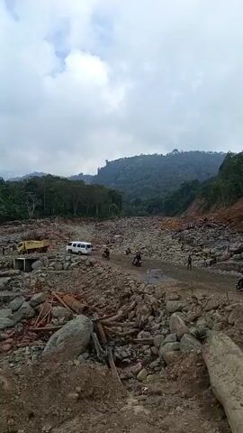 The video shows an area filled with rocks and debris, appearing to be a dried riverbed or a landslide area. There are several vehicles present, including white vans and a yellow truck. People can be seen moving around, indicating some activity in the area. The surrounding environment includes lush green trees and hills, suggesting a natural or rural setting. The sky is overcast, contributing to a somewhat somber or uncertain atmosphere. The overall scene suggests a location where some kind of event, possibly natural disaster or extensive construction work, has taken place.