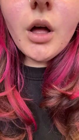 A close-up video shows the lower half of a person's face, featuring their mouth, chin, and part of their neck, set against a dark top. The person has light skin with some small blemishes or freckles, and their hair, visible on either side of their face, is a vibrant pink mixed with darker brown tones, styled with waves. A nose ring is faintly visible at the top edge of the frame.

The activity involves the person blowing smoke rings. They open their mouth, exhale a puff of smoke, and then skillfully form a clear, circular smoke ring that floats upwards. This action is repeated within the short clip. The person's expression is neutral, concentrating on the task. The setting appears to be indoors, though no specific background details are discernible. This event takes place in Beer Yaakov, Israel.