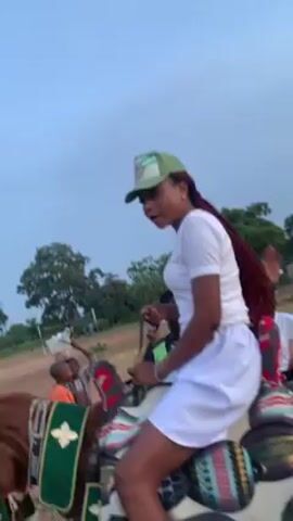 Here is a description of the video:

A woman with red braids rides a brown horse in a dirt field. She is wearing a white t-shirt and shorts, a green hat, and white sneakers. The horse is adorned with colorful fabric and tassels. Several people stand around watching her. The sky is overcast. Trees and buildings are visible in the background.