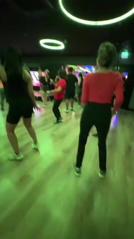 A dynamic indoor scene shows a group of people, predominantly women, engaged in a fitness or dance class in San Isidro, Peru. The participants are actively moving their bodies, following what appears to be a choreographed routine, likely to music (though no audio is transcribed).

The room features a dark ceiling adorned with several bright, circular green LED lights, casting a distinct green hue across the wooden floor. In the background, large screens display vibrant, colorful graphics and visuals, suggesting an interactive fitness experience or an engaging backdrop for the activity. The overall ambiance is energetic and modern, resembling a studio or a club setting. The video exhibits motion blur, indicating the subjects' vigorous movements and possibly the camera's motion. No specific text is visible within the frame.