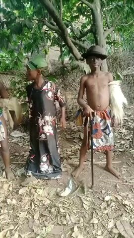 Three children are standing in a wooded area covered in leaves. Two are shirtless and wearing patterned cloth around their waists, and the third is wearing a long floral dress. The child on the right is wearing a hat, holding a walking stick, and has a white, fluffy object attached to their arm. The child on the left is holding a spear-like object. The children are posing for the camera, making hand gestures. The setting appears to be a rural or natural environment.