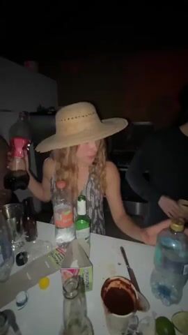 A young woman with blonde, curly hair and a wide-brimmed straw hat is holding a partially full bottle of Coca-Cola in her left hand. She is wearing a dark and light patterned dress. She is looking to her right and appears to be speaking or singing, with her mouth open. Her right hand is gesturing towards the viewer with an open palm. In front of her, on a white table, are several other bottles, including a bottle of water with an orange cap and a green bottle. The background is dark and out of focus, with some indistinct shapes suggesting an indoor or outdoor setting at night. The lighting is dim and appears to be artificial, with some red tones visible.