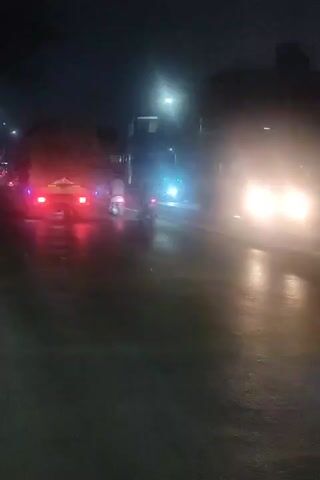 The video shows a nighttime view of traffic on a wet road. Several vehicles are visible, including cars and possibly a truck or bus. Headlights and taillights are illuminated, creating streaks of light due to the motion and the wet surface. Streetlights are also visible in the background. The overall impression is that of a busy road at night, possibly in urban or suburban surroundings, with some of the vehicles appearing to be in motion. The camera appears to be mounted on a moving vehicle, possibly capturing the scene from a driver's perspective or from a passenger's side window.