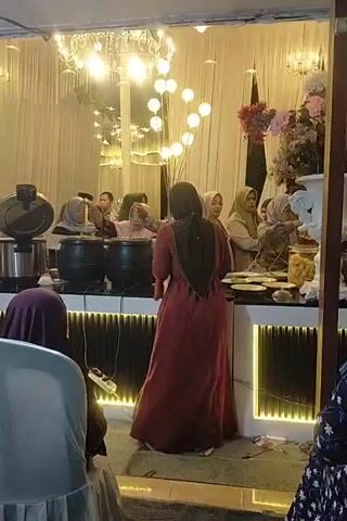 A group of people are gathered around a buffet table. Two large black pots are on the table, and several people are serving themselves food. There are also decorative lights and flowers in the background. The scene appears to be at an indoor event, possibly a wedding reception or party.