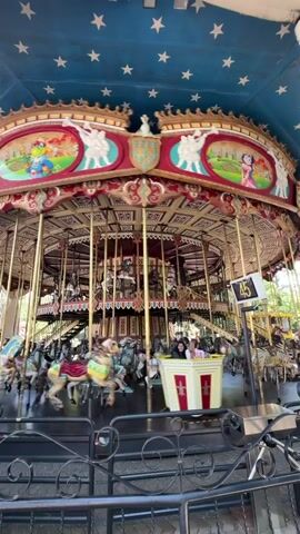 This video shows a carousel in motion. The carousel is decorated with red and gold accents and features a blue ceiling with white stars. The carousel has multiple levels of horses and other animals for riders to sit on, many of which are brightly colored. Some people are visible riding the carousel, and the horses appear to be moving up and down as the carousel rotates. The overall atmosphere suggests a lively amusement park or fairground setting.
