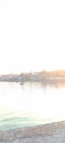 The video shows a body of water with a village in the background. The sun is setting, casting a warm glow over the scene. Several boats are visible on the water, and some houses are built on stilts over the water. The sky is bright and hazy due to the setting sun. The overall impression is one of peace and tranquility.