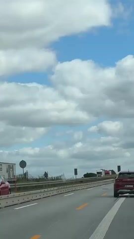 This is a shot from a moving vehicle, looking forward along a highway. The sky is partly cloudy with large white clouds against a blue background. The highway has multiple lanes, separated by a concrete barrier with white vertical elements. Yellow lane markings are visible on the asphalt. Several trucks and cars are driving on the highway, both in the foreground and background. There are also some trees and signs visible beyond the barrier. The camera is tilted slightly upwards, capturing the sky and distant traffic.
