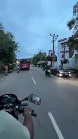 The video depicts a first-person perspective from someone riding a motorcycle on a road in a populated area. The road has a white dividing line and is flanked by buildings, trees, and utility poles. A red bus is driving in the same direction as the motorcycle. Other vehicles, including a tuk-tuk and a car, are visible along the road. The sky is overcast, suggesting it might be dusk or early morning. The rider's arm and hand can be seen holding the motorcycle handlebars.