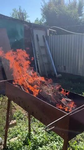 A barbecue grill with wood burning in it. The flames are tall and orange. In the background there is a fence and some trees. The sky is blue.