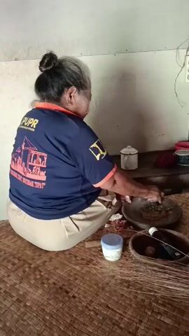 An elderly woman with gray hair is squatting on a woven mat floor, facing a wall, and grinding spices in a stone mortar with a pestle. She is wearing a dark blue t-shirt with orange trim and khaki pants. The t-shirt has an emblem on the back featuring the letters "PUPR" and a depiction of building construction. Beside her are several containers and bowls on the floor and on a shelf behind her. An electrical outlet is visible on the wall.