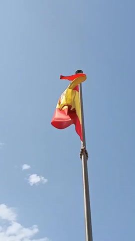 A Spanish flag waves in the wind against a clear blue sky with a few wispy clouds. The flag is attached to a metal flagpole.