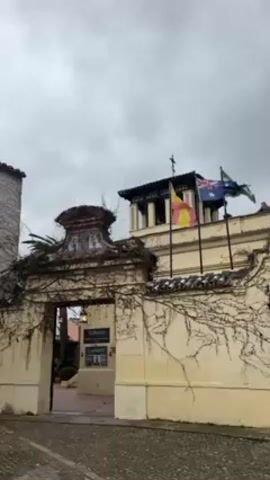 The video shows an exterior shot of a building on a cloudy day. The building has a stone tower and a wall with a gate. The wall is covered in vines. Three flags are flying on top of the building: the Spanish flag, the Australian flag, and the flag of Andalusia. A sign hangs inside the gate. The ground is paved with cobblestones. A person walks across the paved area.