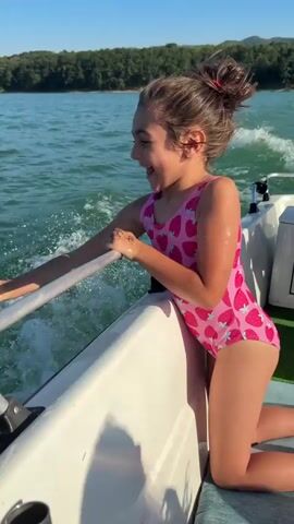 A young girl with her hair in a ponytail, wearing a pink swimsuit with a strawberry print, is kneeling on the side of a boat. She is holding onto a metal railing. The boat is moving through clear, green water, creating a wake. The girl has a wide, excited expression, with her mouth open and eyes looking to the left. Her right arm is extended, and her left hand is gripping the railing. Sunlight is reflecting off the water, and the sky is a clear blue. In the background, there is a line of trees on a distant shore, suggesting a natural landscape. The time of day appears to be sunny and warm, likely afternoon. The overall scene conveys a sense of joy and excitement, possibly during a boat ride on a lake or river.