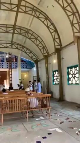 The video shows the interior of a church or gathering place with several people standing in front of a sign that reads "Bukit Rhema". The building has a high, arched ceiling with a metal framework. There are rows of wooden pews facing the sign. People are standing near the sign, and some are talking and interacting. A man is sitting in one of the pews near the front. The floor is decorated with colorful geometric patterns. The overall setting suggests a religious or community gathering place.