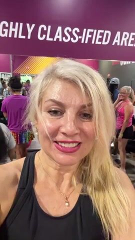 A group of women are in a gym. A blonde woman in the foreground is filming herself. She is smiling and wearing a black tank top and a necklace. Behind her, women are exercising on yoga mats. There is a sign in the background that reads "HIGHLY CLASSIFIED AREA".