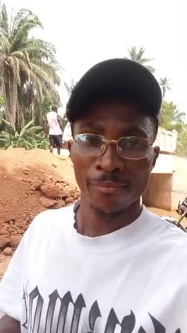 The video opens with a close-up selfie of a Black man in a white t-shirt, black cap, and gold-rimmed glasses. He has a mustache and goatee, wears a gold watch on his left wrist, and a ring on his right hand, which he often rests on his chin.

The setting is an outdoor, sunny, and dusty environment in what appears to be Aloko, Nigeria. Behind the man are large piles of reddish-brown dirt and rocks. In the initial frames, a white dump truck is visible in the background. As the camera pans slightly, a group of other Black men appears, some shirtless, others in t-shirts or a tank top with "CHICAGO" visible on it. A red and black motorbike is also seen parked nearby.

Further into the video, the dump truck is shown on an elevated dirt path or bridge, with a concrete culvert or underpass beneath it. The background also features numerous palm trees and lush green vegetation, indicative of a tropical climate. The ground is consistently composed of dirt and sand.

The man in the foreground maintains a thoughtful or neutral expression, occasionally panning the camera to reveal more of his surroundings. The overall scene suggests a construction or roadwork site, possibly involving earthmoving activities, with people gathered around under clear, bright daytime skies.