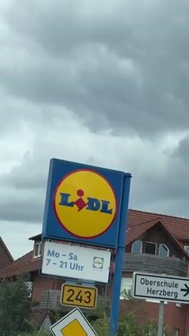 A close-up, low-angle shot of a Lidl supermarket sign. The sign is blue with a large yellow circle in the center. Inside the circle is the Lidl logo, which consists of the letters "LIDL" in blue with a red abstract figure in between the "L"s. The background is a cloudy sky.