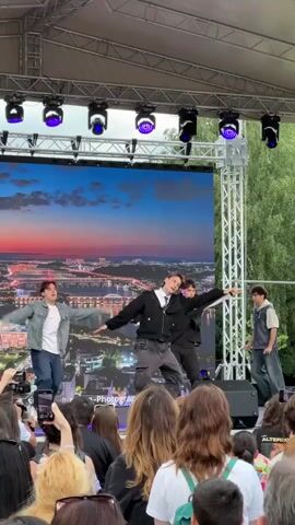 A group of five young men are performing a synchronized dance routine on an outdoor stage in front of a large LED screen displaying a cityscape at sunset. The men are dressed in casual, stylish clothing. From left to right, the first performer is wearing a denim jacket, a white t-shirt, and blue jeans. The second performer is in a black hoodie and dark pants. The third performer is wearing a black jacket, a white shirt, and dark pants. The fourth performer, in the center, is wearing a dark, loose-fitting shirt and dark pants. The fifth performer on the right is wearing a white shirt and dark pants. The stage is covered by a white canopy and equipped with professional lighting. A crowd of people, mostly young women, are visible in the foreground, many holding up their phones to record the performance. The background shows trees and greenery, suggesting the performance is taking place in a park or outdoor venue. The overall atmosphere appears to be energetic and lively.
