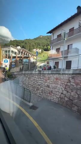 The video shows a view from a moving vehicle, likely a bus, looking out a window. The scene is an outdoor environment with a clear blue sky and sunshine. In the foreground, the window has a reflective surface showing streaks and distortions. A yellow line marks the edge of a road. A metal railing runs along the side of the road. A stone wall with rough-hewn rocks is visible beyond the railing. Above the wall, there are buildings with balconies and signs, indicating a town or village. A traffic sign for a roundabout is visible. In the background, a green, tree-covered mountainside rises sharply, with a ski lift or cable car system visible ascending the slope. Some people are walking on a path behind the railing. The overall impression is a scenic view of a mountain town.