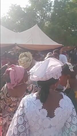 The video captures a scene at an outdoor wedding, with guests dressed in colorful attire. A man in a black robe and baseball cap engages in conversation with a woman wearing a head wrap. A red carpet is visible. The background features tents, trees, and the outline of buildings. Music plays in the background. The video appears to be shot with a handheld camera.