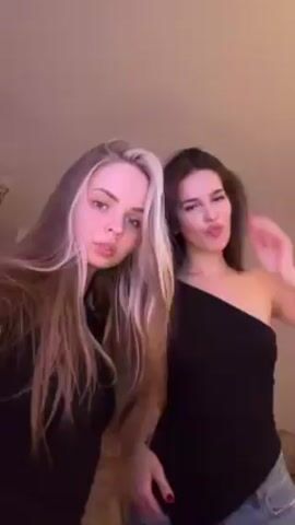 Two young women pose for a video. The woman on the left has long brown hair with a blonde streak and wears a black shirt and jeans. The woman on the right has long dark brown hair and wears a one-shoulder black top and jeans. They are standing indoors against a plain background with a ceiling lamp visible in the upper left corner. They make playful faces and slight hand gestures. The lighting is soft and warm, with a slight reddish tint.