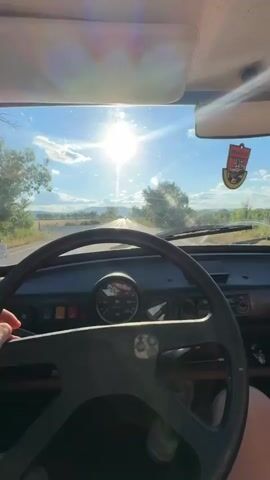 The video shows a first-person view from inside a car. The sun is shining brightly, creating a lens flare effect. The driver's hand is visible on the steering wheel. The dashboard of the car is visible, including the speedometer. The car is on a paved road, with fields and trees on either side. The sky is blue with some clouds. A sticker is visible on the passenger side sun visor. The video appears to be filmed while the car is in motion.