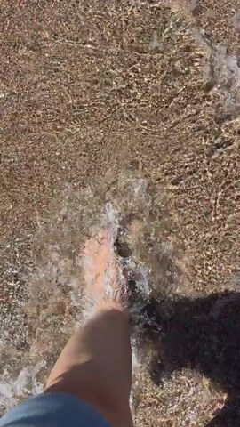 The video shows a first-person perspective of someone walking into shallow water. The water is clear and calm, with sunlight reflecting off the surface. The ground beneath the water appears to be composed of small pebbles and sand. A shadow of a person's legs and feet is visible on the right side of the frame, indicating they are moving forward into the water. The person's left foot is submerged in the water and appears to be the focus of the shot. The overall impression is one of a peaceful and serene environment, likely a beach or shallow body of water.